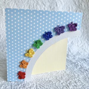 A hand cut, blue spotty card with 7 individually crocheted and hand sewn butterflies in the colours of the rainbow, arranged in an arch over a spotty yellow sun. This card has a blank white paper insert for you to write your own message. Envelope included. Approximate size 15cm x 15cm. Handmade, using 100% cotton.