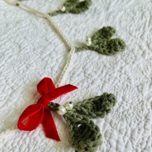 A handmade and crocheted garland of 9 green mistletoe and pearl berry sprigs, with a red velvet bow detail. Made using 100% cotton and glass beads.