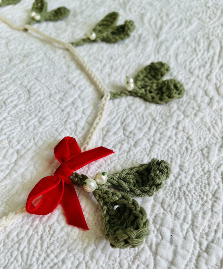 A handmade and crocheted garland of 9 green mistletoe and pearl berry sprigs, with a red velvet bow detail. Made using 100% cotton and glass beads.
