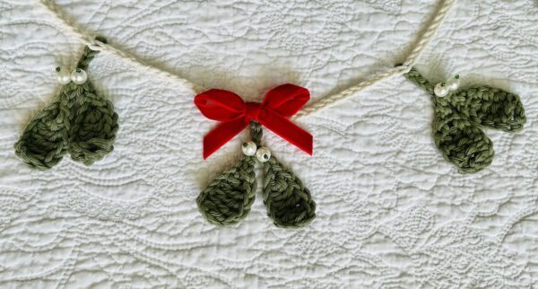 A handmade and crocheted garland of 9 green mistletoe and pearl berry sprigs, with a red velvet bow detail. Made using 100% cotton and glass beads.