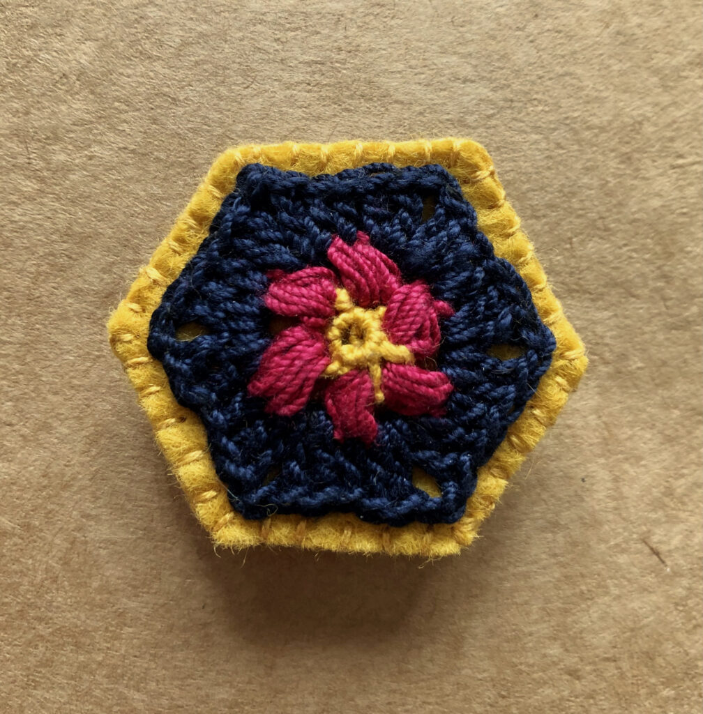 A hexagon shaped, hand stitched, felt and flower design crocheted brooch with locking brooch fixing.