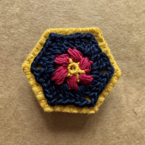 A hexagon shaped, hand stitched, felt and flower design crocheted brooch with locking brooch fixing.