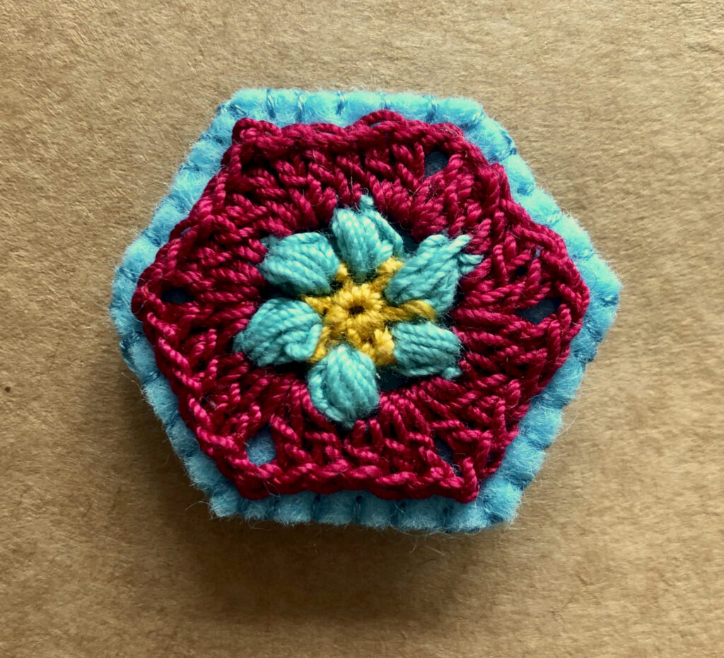 A hexagon shaped, hand stitched, felt and flower design crocheted brooch with locking brooch fixing.