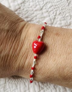 A red ceramic heart and red and clear glass beaded bracelet on a cream crocheted cotton strap, with a fully adjustable sliding fastening.