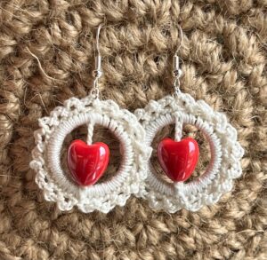 A handmade, crocheted pair of ivory coloured and red heart beaded hoop earrings, made using 100% cotton and ceramic heart beads with metal fixings. The earring hooks are made from a silver plated brass and are nickel, lead and cadmium free. Each earring measures approximately 3.4cm wide by 5cm Height (including hook). No plastic has been used. This item is Vegan and Eco-friendly, reusable and fully recyclable. 