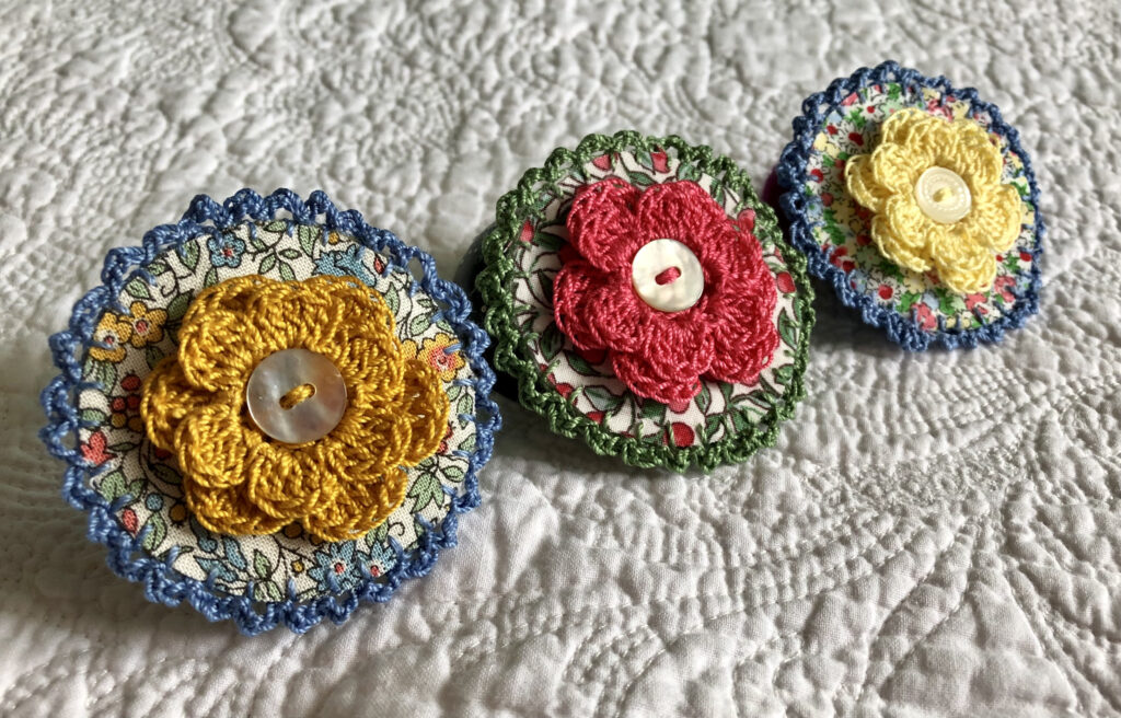 A round brooch made using a patterned fabric background with a crocheted flower in the centre and  crocheted edging. Handmade using 100% cotton fabric and yarn. With button detail and a locking brooch fastening on a felt backing.