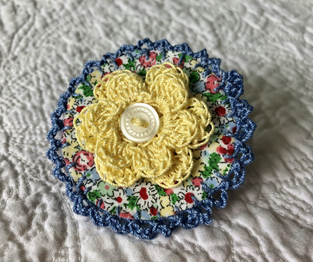 A round brooch made using a patterned fabric background with a crocheted flower in the centre and crocheted edging. Handmade using 100% cotton fabric and yarn. With button detail and a locking brooch fastening on a felt backing.