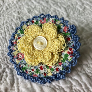 A round brooch made using a patterned fabric background with a crocheted flower in the centre and crocheted edging. Handmade using 100% cotton fabric and yarn. With button detail and a locking brooch fastening on a felt backing.