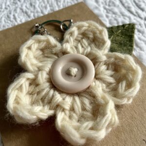 A hand crocheted cream coloured flower, with a hand embroidered green felt leaf and a button detail on a metal keyring/bag charm clip.