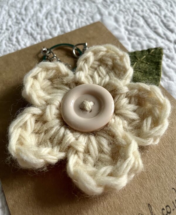 A hand crocheted cream coloured flower, with a hand embroidered green felt leaf and a button detail on a metal keyring/bag charm clip.