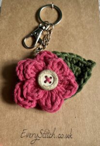 A hand crocheted, pink coloured layered flower with green leaf and a wooden button detail on a metal keyring/bag charm clip.