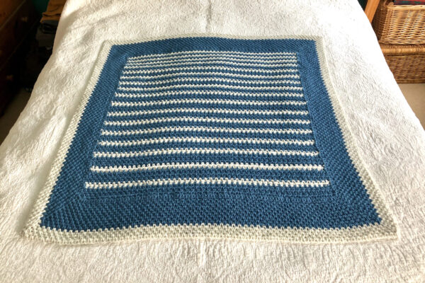 A handmade, crocheted blanket in light denim blue and light cream. Made with 80%Acrylic/20% Wool mix yarn. Approximate size 92cm Width x 92cm Height.