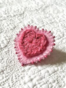 A single crocheted heart in a heathered raspberry colour, mounted on a hand stitched pink felt heart and decorative, crocheted cotton edging and glass beaded embellishments. The fixing is a locking metal brooch fastening on the back. Approximate size 5cm width x 5cm height.