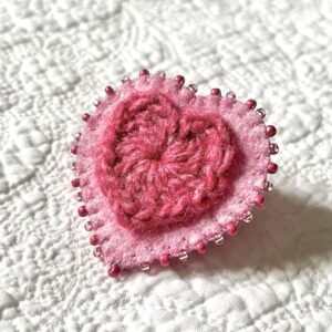 A single crocheted heart in a heathered raspberry colour, mounted on a hand stitched pink felt heart and decorative, crocheted cotton edging and glass beaded embellishments. The fixing is a locking metal brooch fastening on the back. Approximate size 5cm width x 5cm height.