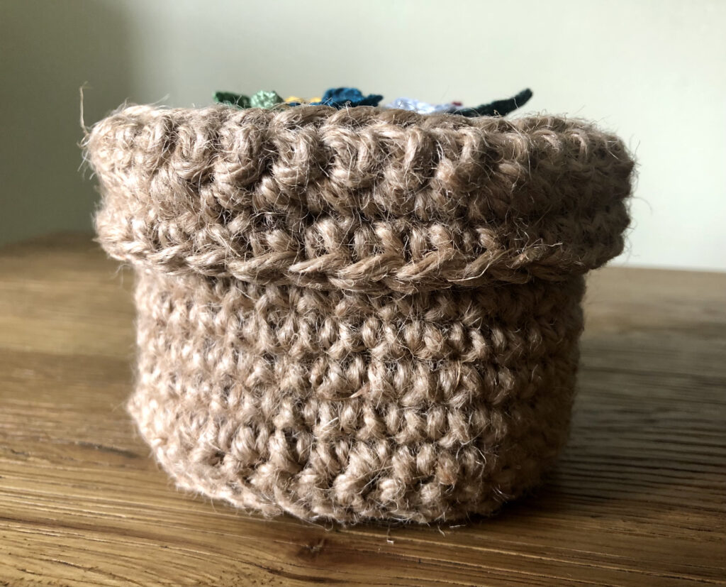 A handmade, crocheted 100% jute basket with lid. Decorated with 100% cotton crocheted flowers and leaves. Approximate size 7.5cm Height x 11.5cm width.