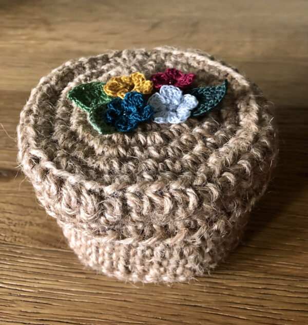 A handmade, crocheted 100% jute basket with lid. Decorated with 100% cotton crocheted flowers and leaves. Approximate size 7.5cm Height x 11.5cm width.