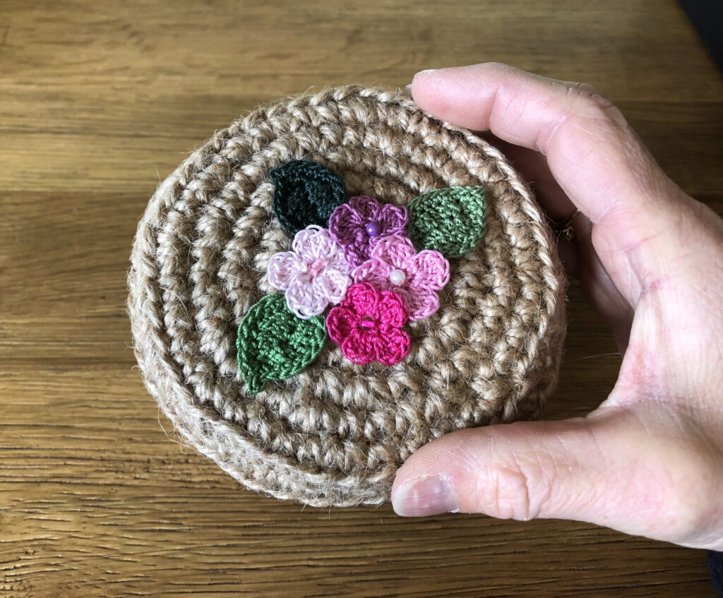 A handmade, crocheted 100% jute basket with lid. Decorated with 100% cotton crocheted flowers and leaves. Approximate size 7.5cm Height x 11.5cm width.