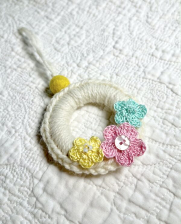 A mini crocheted and embellished Spring/Easter inspired flower wreath. Handmade using a wooden ring with a mix of acrylic, cotton and wool materials, glass beads and recycled buttons.