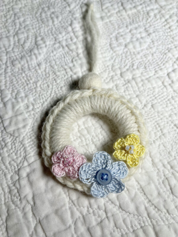 A mini crocheted and embellished Spring/Easter inspired flower wreath. Handmade using a wooden ring with a mix of acrylic, cotton and wool materials, glass beads and recycled buttons.