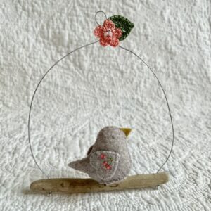 A single small sized bird, handmade in beige felt, with a cotton multi coloured flower print fabric chest and hand embroidered/glass beaded detail on the wings. This bird is sat on a natural driftwood perch with a wire hanger that is decorated with a crocheted pink flower and green leaf detail.