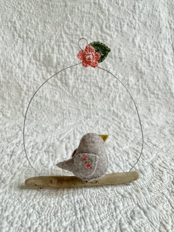 A single small sized bird, handmade in beige felt, with a cotton multi coloured flower print fabric chest and hand embroidered/glass beaded detail on the wings. This bird is sat on a natural driftwood perch with a wire hanger that is decorated with a crocheted pink flower and green leaf detail.