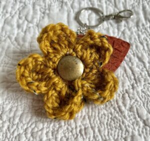 A hand crocheted Mustard coloured flower, with a hand embroidered rust coloured felt leaf and a brass button detail on a metal keyring/bag charm clip.