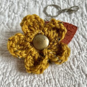 A hand crocheted Mustard coloured flower, with a hand embroidered rust coloured felt leaf and a brass button detail on a metal keyring/bag charm clip.