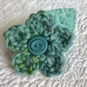 A handmade brooch, crocheted wool flower in greens with a hand embroidered leaf and wooden button detail.