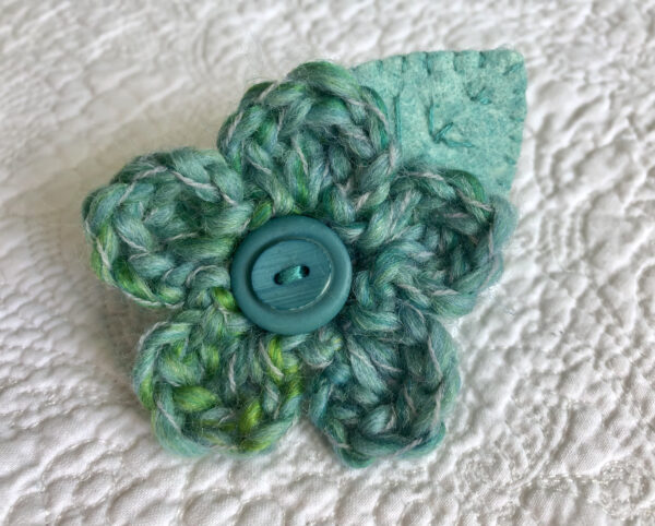 A handmade brooch, crocheted wool flower in greens with a hand embroidered leaf and wooden button detail.