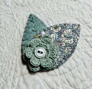 A single, double layered, crocheted flower in duck egg colour, with vintage button detail and two leaf brooch. Flower made using 100% Cotton with hand stitched felt and patterned fabric leaves. A locking metal brooch fastening on the back. Approximate size 7.5cm width x 6.5cm height.