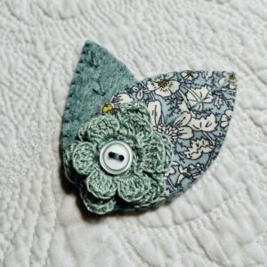 A single, double layered, crocheted flower in duck egg colour, with vintage button detail and two leaf brooch. Flower made using 100% Cotton with hand stitched felt and patterned fabric leaves. A locking metal brooch fastening on the back. Approximate size 7.5cm width x 6.5cm height.