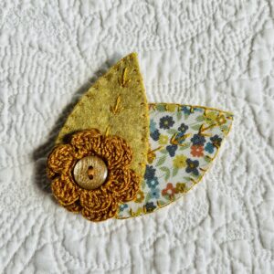 A single, double layered, crocheted flower in a mustard colour, with vintage button detail and two leaf brooch. Flower made using 100% Cotton with hand stitched felt and patterned fabric leaves. A locking metal brooch fastening on the back.