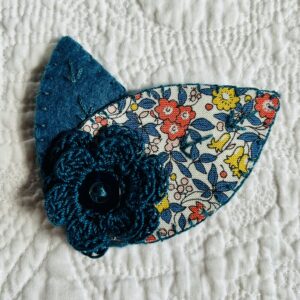 A single, double layered, crocheted flower in a teal colour, with vintage button detail and two leaf brooch. Flower made using 100% Cotton with hand stitched felt and patterned fabric leaves. A locking metal brooch fastening on the back. Approximate size 7.5cm width x 6.5cm height.