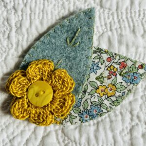 A single, double layered, crocheted flower in a mustard colour, with vintage button detail and two leaf brooch. Flower made using 100% Cotton with hand stitched felt and patterned fabric leaves. A locking metal brooch fastening on the back. Approximate size 7.5cm width x 6.5cm height.