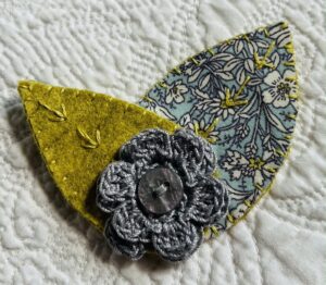 A single, double layered, crocheted flower in a light grey colour, with vintage button detail and two leaf brooch. Flower made using 100% Cotton with hand stitched felt and patterned fabric leaves. A locking metal brooch fastening on the back. Approximate size 7.5cm width x 6.5cm height.