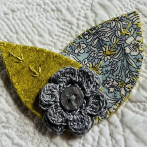 A single, double layered, crocheted flower in a light grey colour, with vintage button detail and two leaf brooch. Flower made using 100% Cotton with hand stitched felt and patterned fabric leaves. A locking metal brooch fastening on the back. Approximate size 7.5cm width x 6.5cm height.