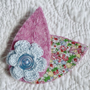 A single, double layered, crocheted flower in a light blue colour, with vintage button detail and two leaf brooch. Flower made using 100% Cotton with hand stitched felt and patterned fabric leaves. A locking metal brooch fastening on the back. Approximate size 7.5cm width x 6.5cm height.