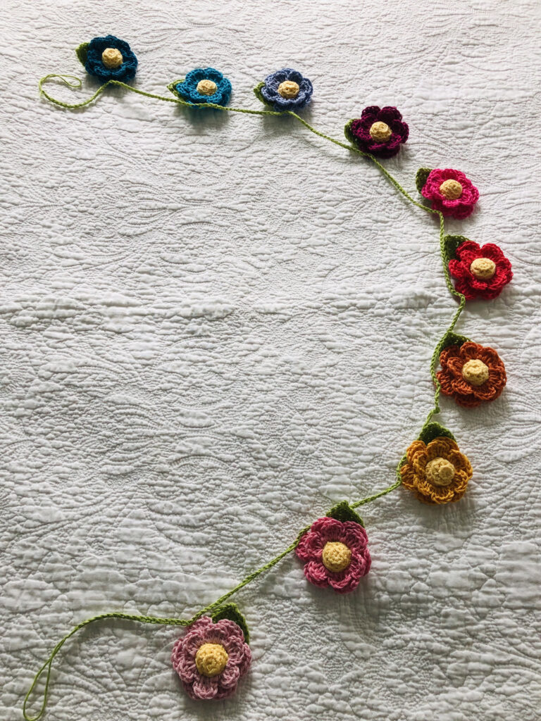 A hand crocheted garland of double layered flowers with crocheted centre and leaf details. Made in a rainbow of colours using 100% cotton.