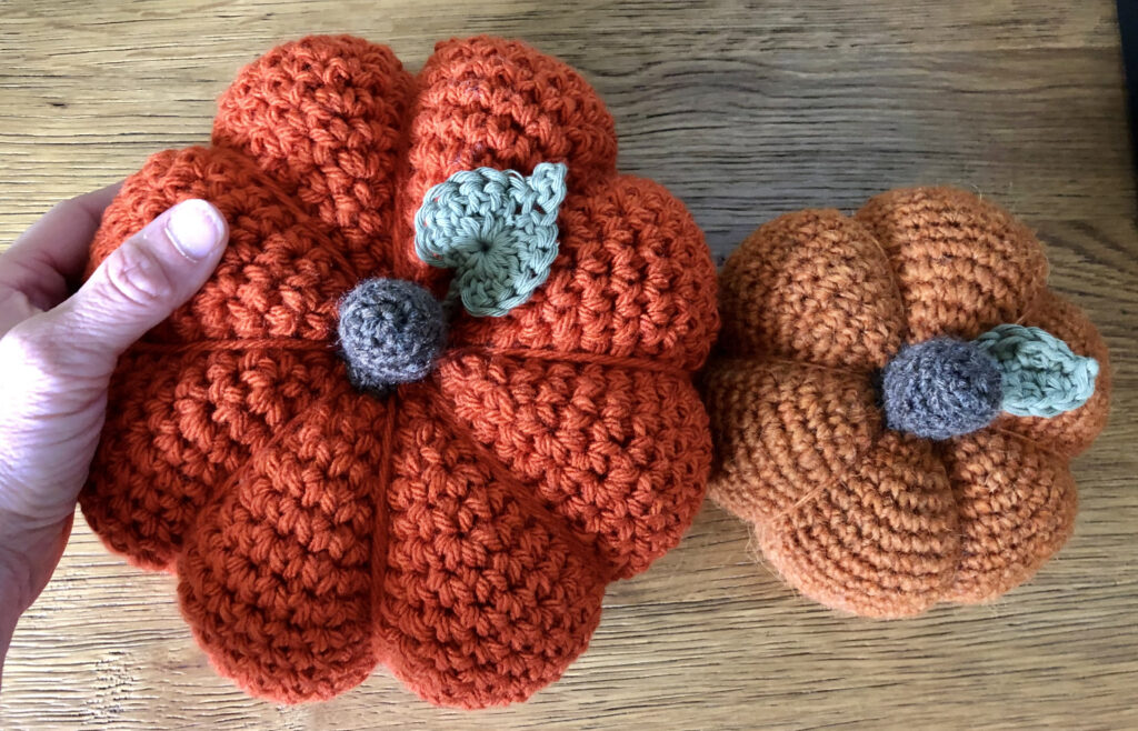 Crochet pumpkins in a selection of sizes, thickness and texture of yarn or wool. In warm tones of orange with a brown stalk and green leaf detail.