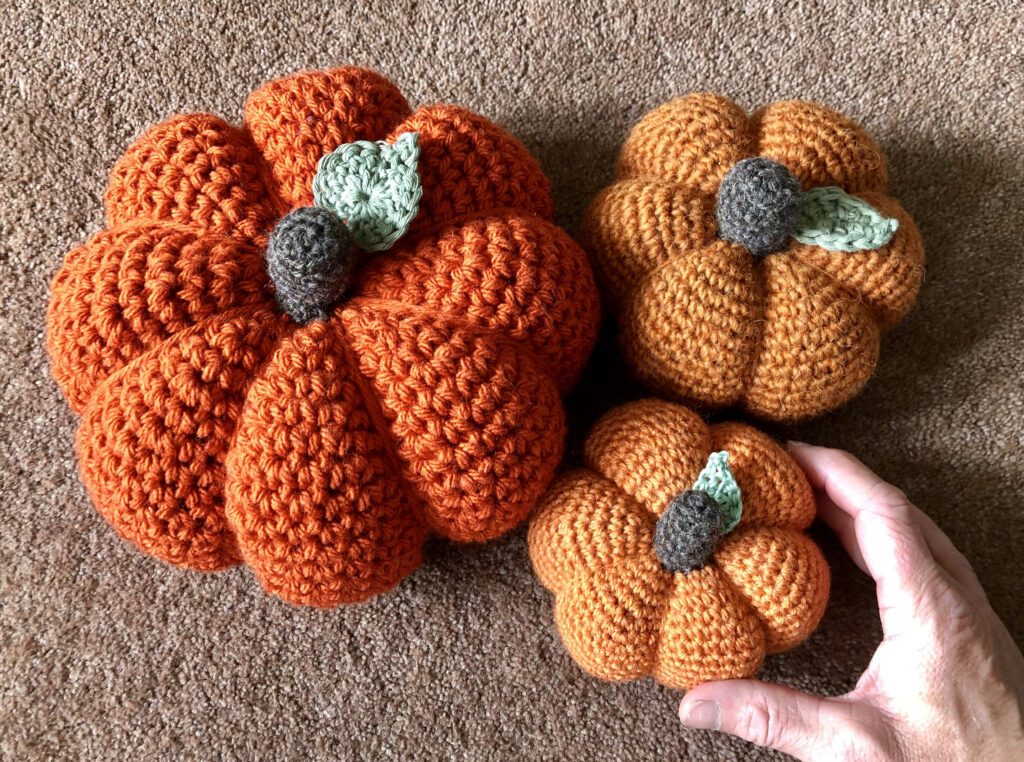 Crochet pumpkins in a selection of sizes, thickness and texture of yarn or wool. In warm tones of orange with a brown stalk and green leaf detail.