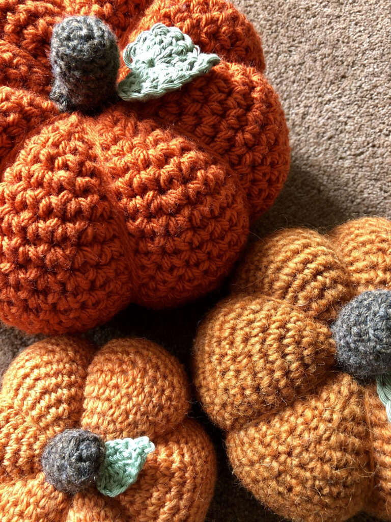Crochet pumpkins in a selection of sizes, thickness and texture of yarn or wool. In warm tones of orange with a brown stalk and green leaf detail.