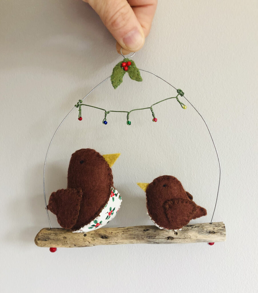 A pair of handmade little birds made with brown felt and a holly print fabric chest. These birds are sat on a natural driftwood perch with a wire hanger that is decorated with felt and beaded holly and wire and beaded christmas lights effect bunting.