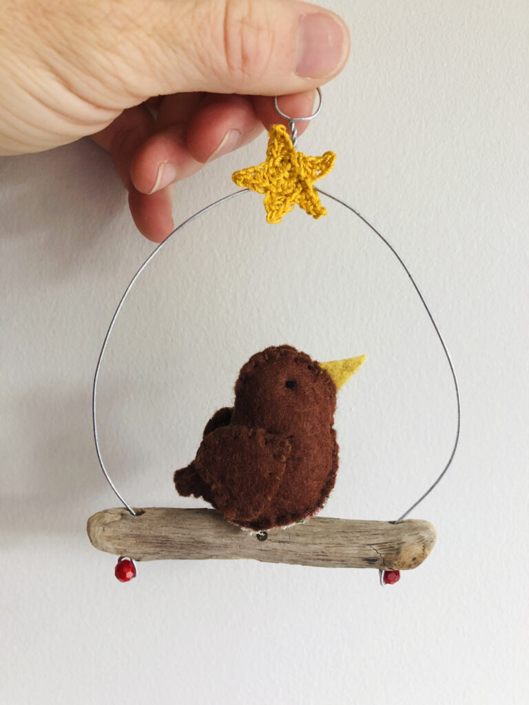 A single, small sized, handmade little robin, made in brown felt with a cotton holly print fabric chest. This bird is sat on a natural driftwood perch with a wire hanger that is decorated with a crocheted yellow star.