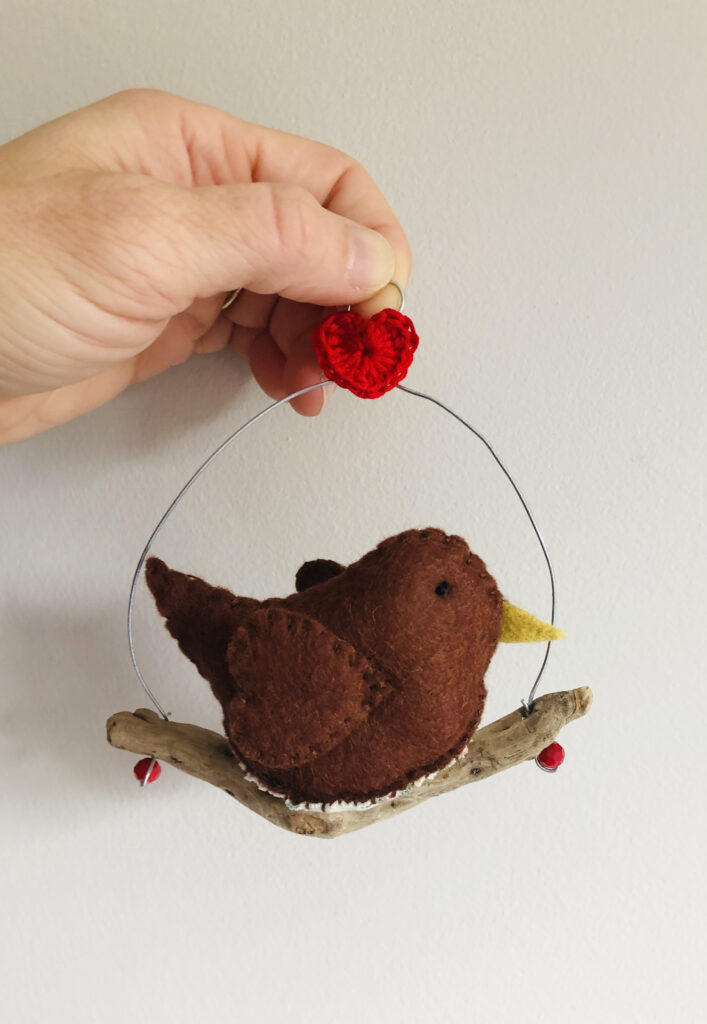 A single, medium sized, handmade little robin, made in brown felt with a cotton holly print fabric chest. This bird is sat on a natural driftwood perch with a wire hanger that is decorated with a crocheted red heart.