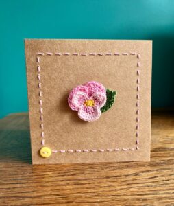 A single crocheted Pink Pansy flower and a green leaf, with hand sewn boarder and button detail. This card has a blank white paper insert for you to write your own message. Handmade, using 100% cotton.