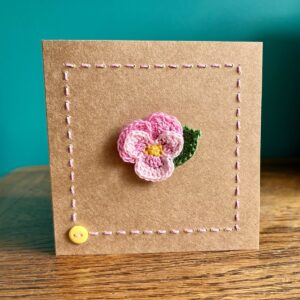 A single crocheted Pink Pansy flower and a green leaf, with hand sewn boarder and button detail. This card has a blank white paper insert for you to write your own message. Handmade, using 100% cotton.