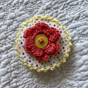 A round brooch made using a patterned fabric background with a crocheted flower in the centre and crocheted edging. Handmade using 100% cotton fabric and yarn. With button detail and a locking brooch fastening on a felt backing.