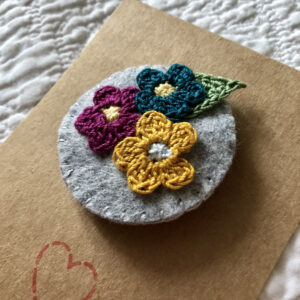 A handmade round brooch made using a felt background with a posy of crocheted flowers and a leaf in the centre. With a metal locking brooch fastening on a felt backing.