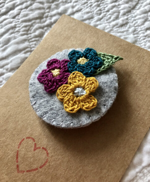 A handmade round brooch made using a felt background with a posy of crocheted flowers and a leaf in the centre. With a metal locking brooch fastening on a felt backing.