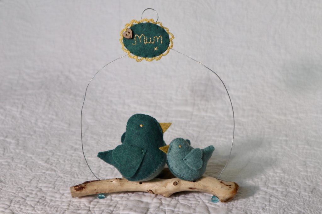 Two teal and green hand sewn felt birds sitting on a driftwood branch with a wire hanging hoop and hand embroidered plaque.